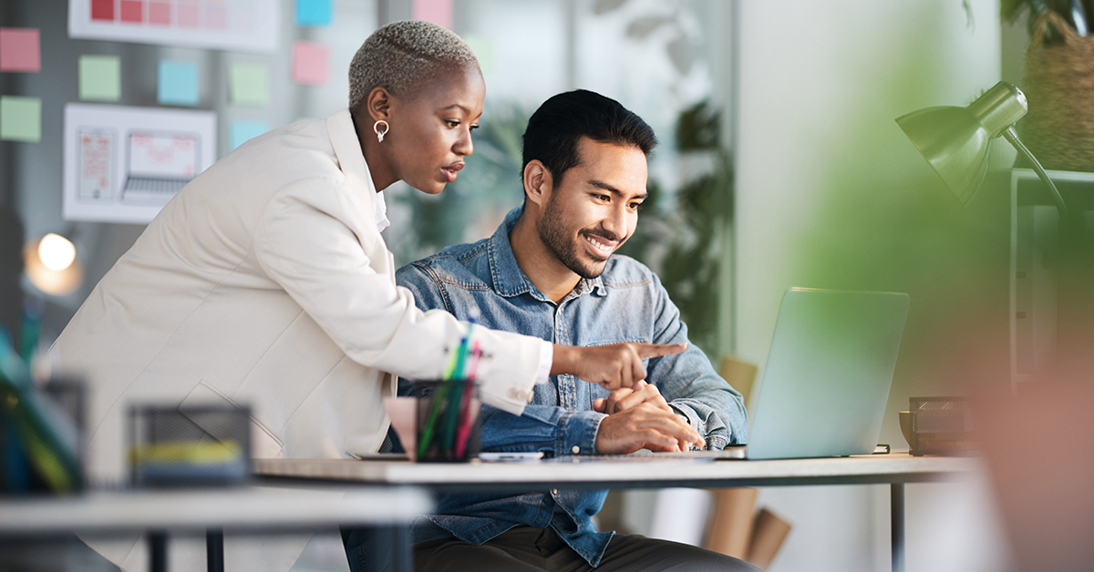woman wearing white coat pointing at computer with man typing