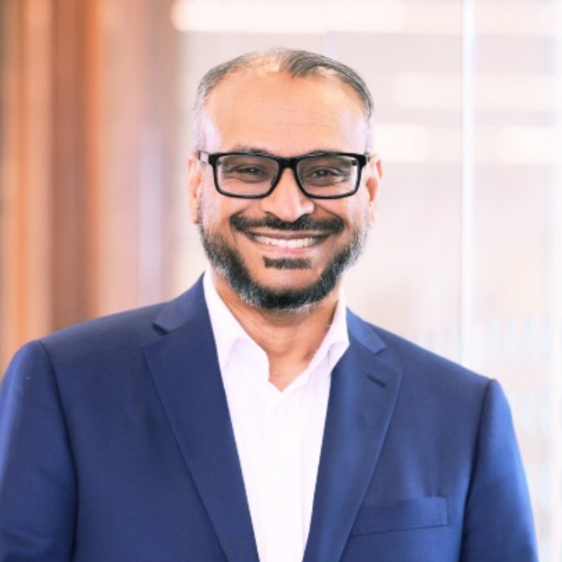 Professional headshot of a man with glasses in a navy blazer against a warm background