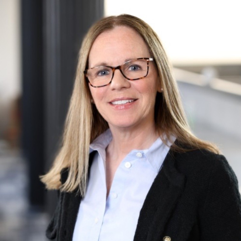 Professional headshot of a woman with long blonde hair and glasses in a black blazer