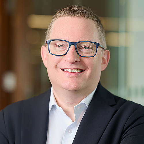 Professional headshot of a man with glasses in a dark blazer