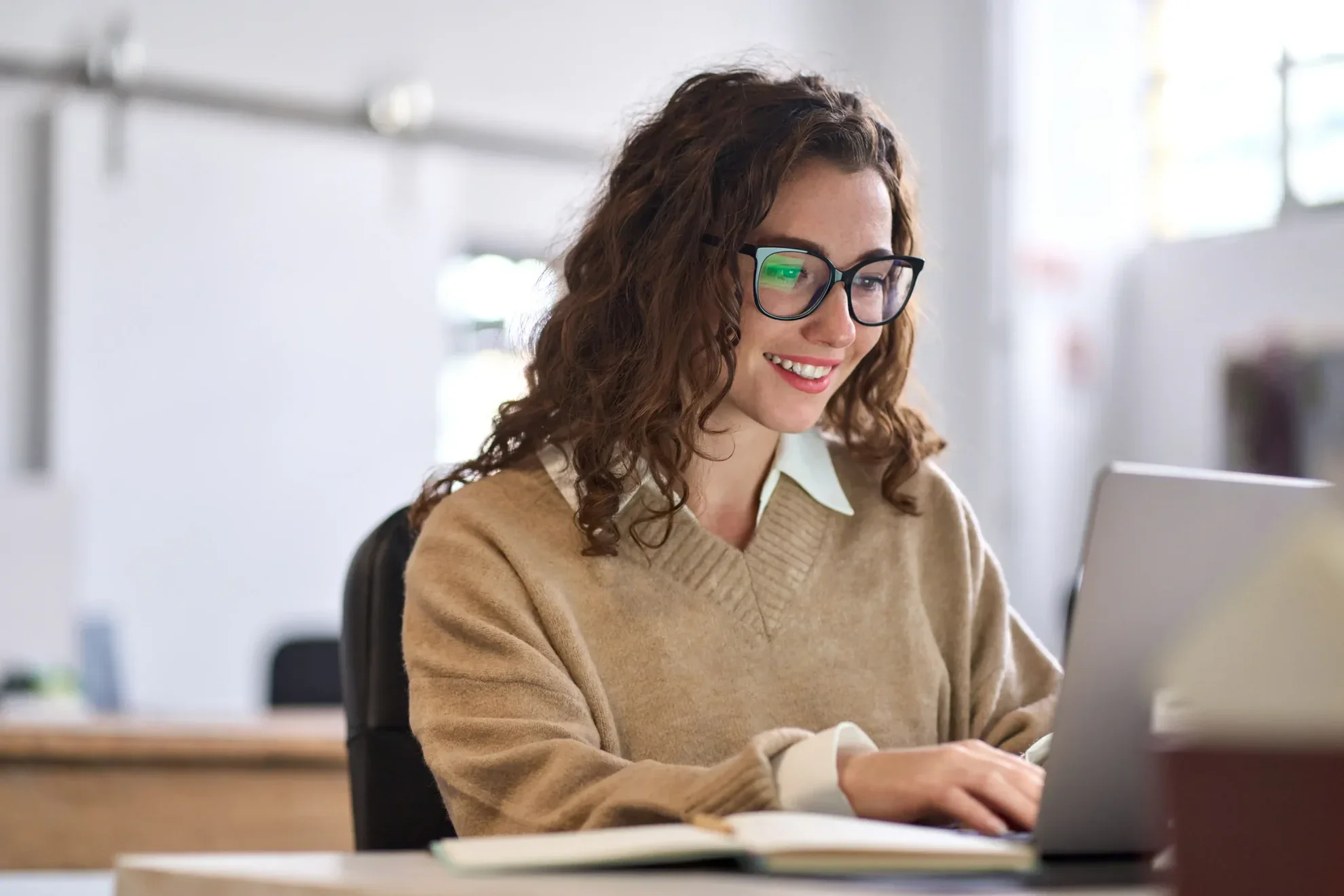 Woman with curly hair and glasses smiling while working on a laptop