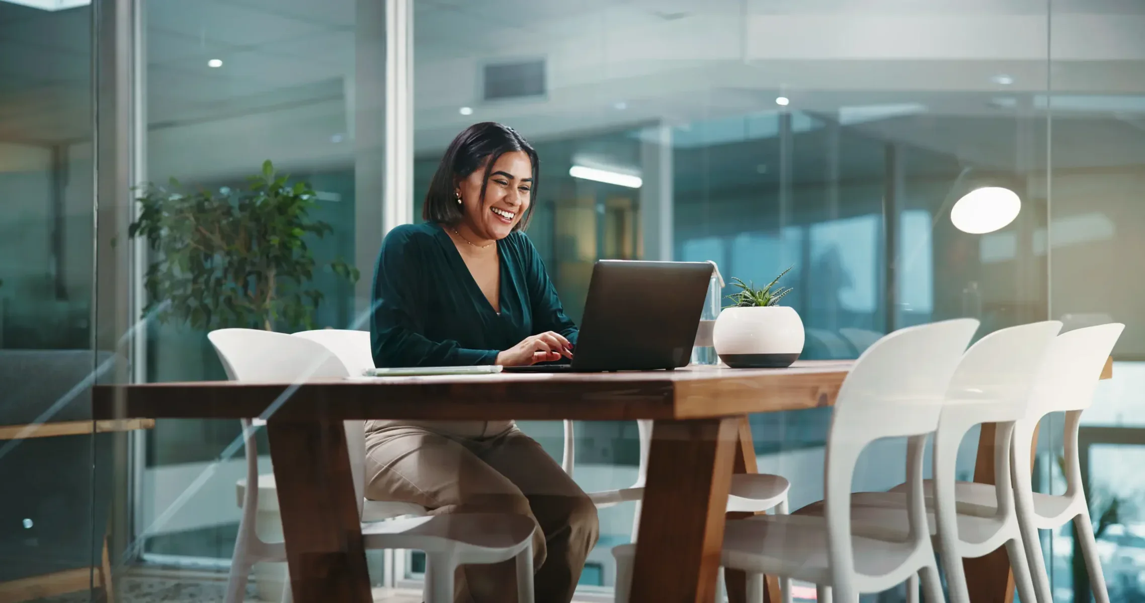 Woman with dark hair seated at a laptop inside a glass-walled modern office