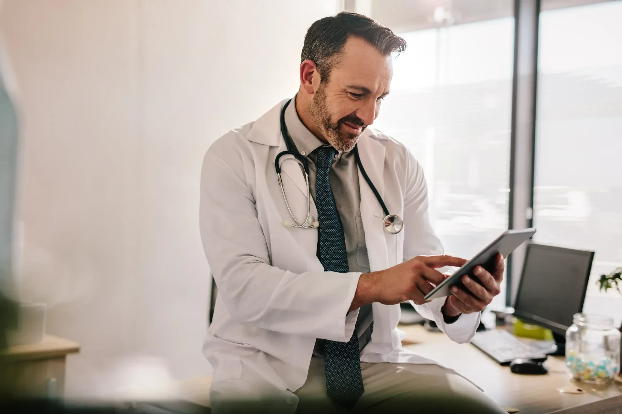 Male doctor in a white coat with a stethoscope reviewing information on a tablet