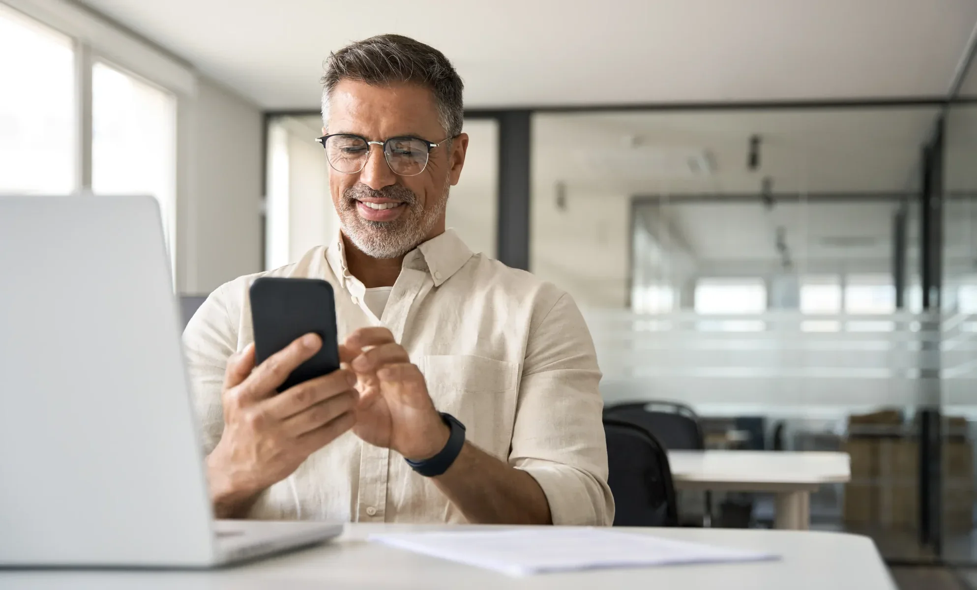 Man with glasses smiling while looking at his smartphone at a desk with a laptop