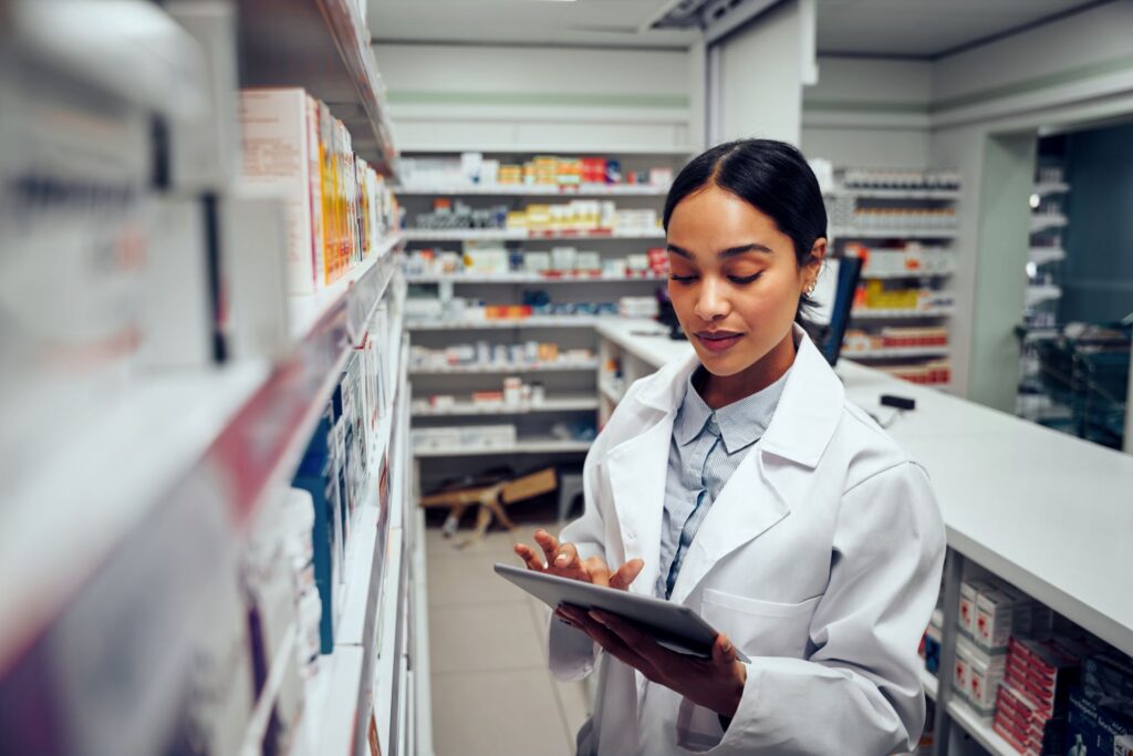 A person in a white lab coat uses a tablet while standing beside shelves stocked with boxed medications in a pharmacy-like setting.