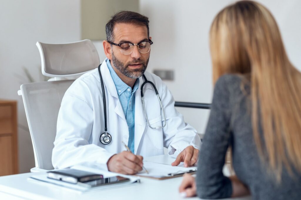 A medical professional wearing a white coat sits at a desk, writing on paperwork while another person sits across the desk during a consultation.