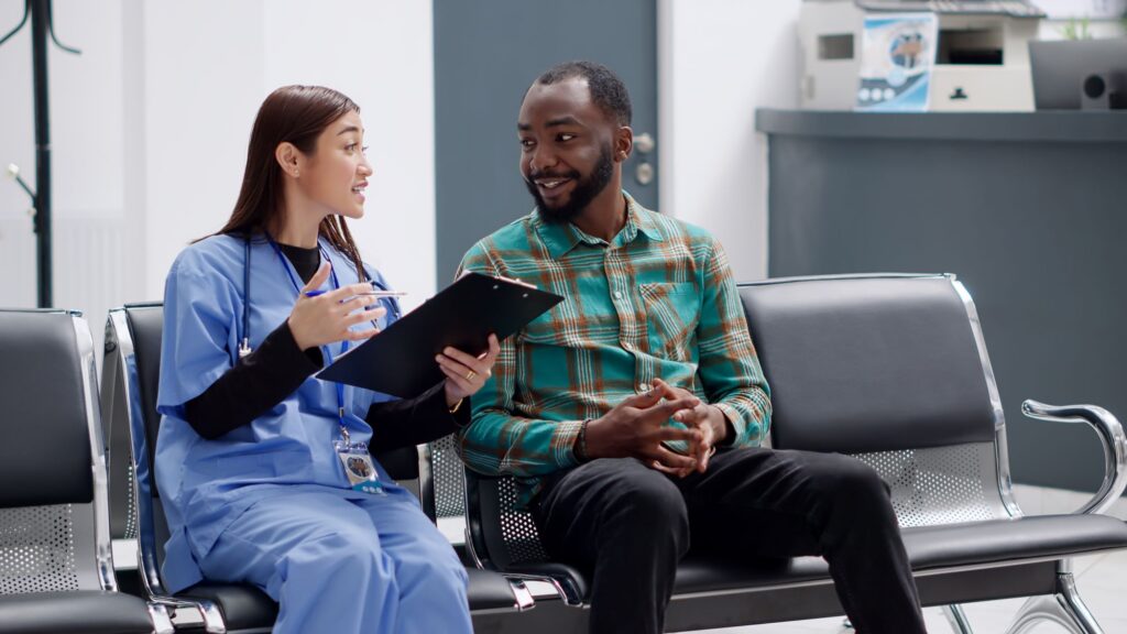 A healthcare worker in medical scrubs sits beside another person in a waiting area, holding a clipboard and discussing information.