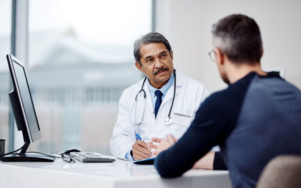 A medical professional in a white coat sits at a desk and speaks with another person across from them, with a computer monitor and paperwork visible.