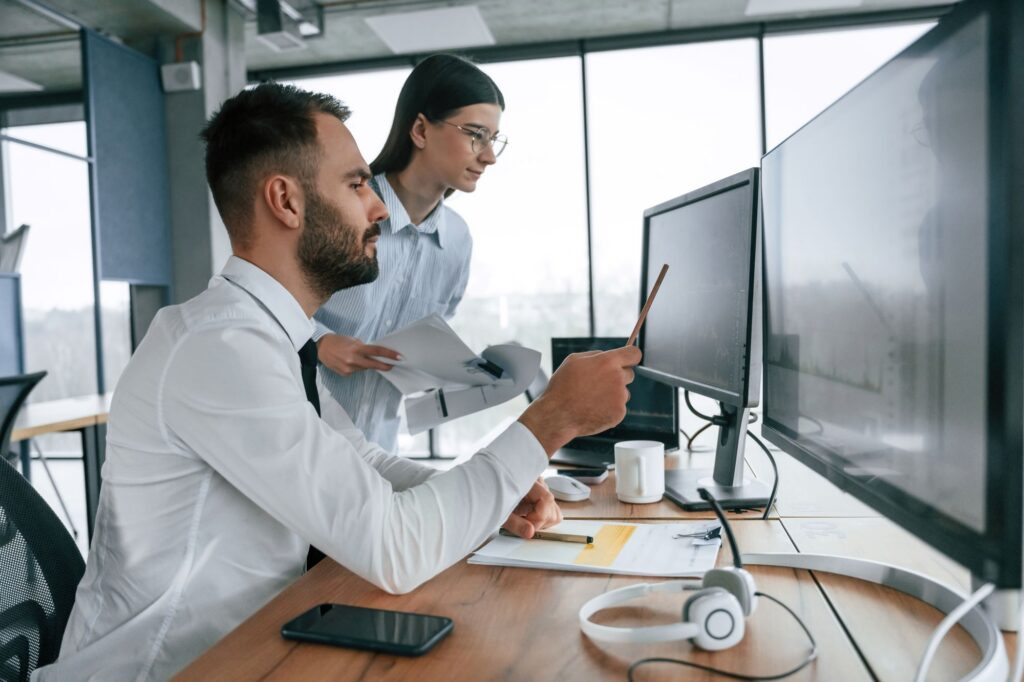 Two people at a desk review information on large computer monitors, with one person pointing at the screen while the other holds printed documents.