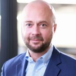 Professional headshot of a bald man with a beard wearing a blue suit jacket