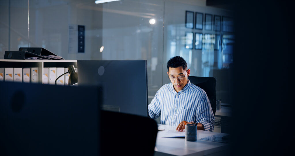 Person wearing a striped shirt reviewing documents at a desk beside a computer monitor in a professional office setting with shelves, binders, and glass walls.