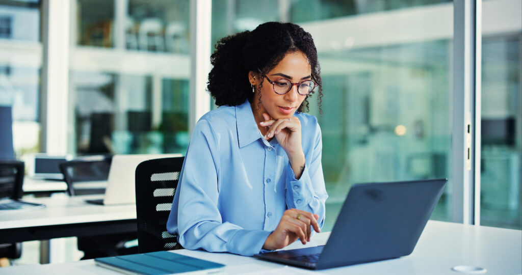 Person wearing glasses and a light blue shirt working on a laptop at a desk in a modern office with large windows and an open workspace in the background.