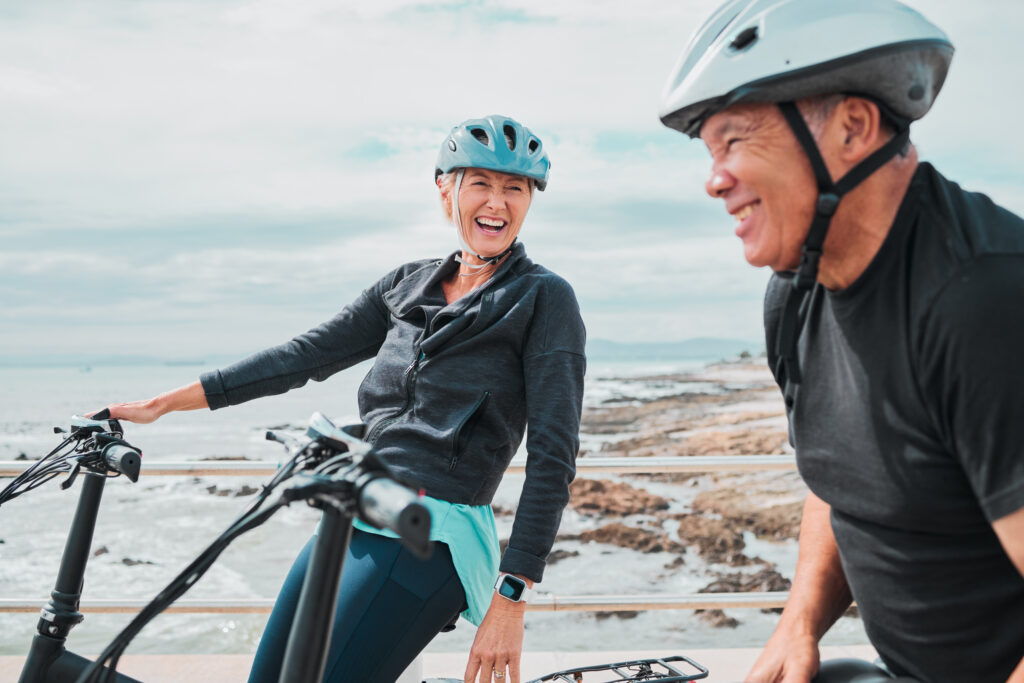 Two older adults wearing bicycle helmets pause with their bikes along a coastal path, with rocky shoreline and ocean visible in the background.